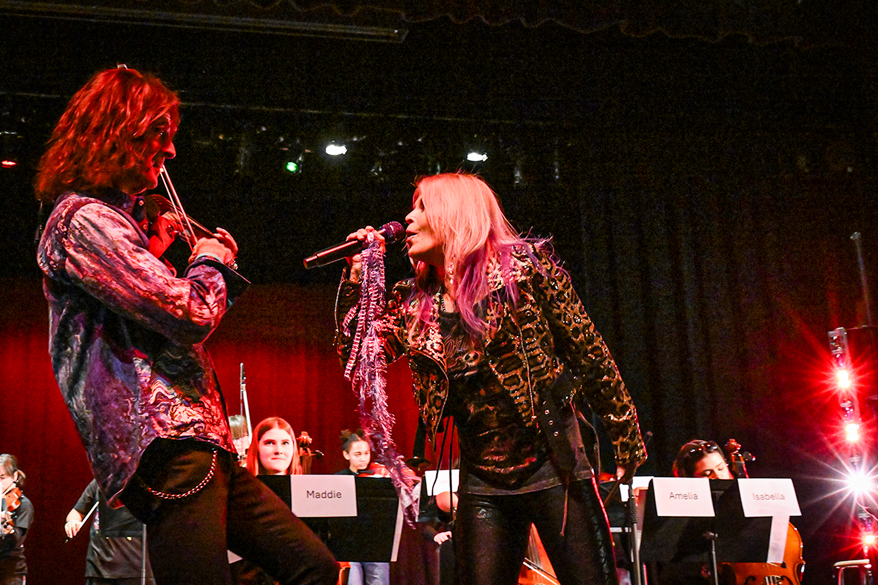 Mark Wood plays violin while a woman sings at him on stage with students behind them