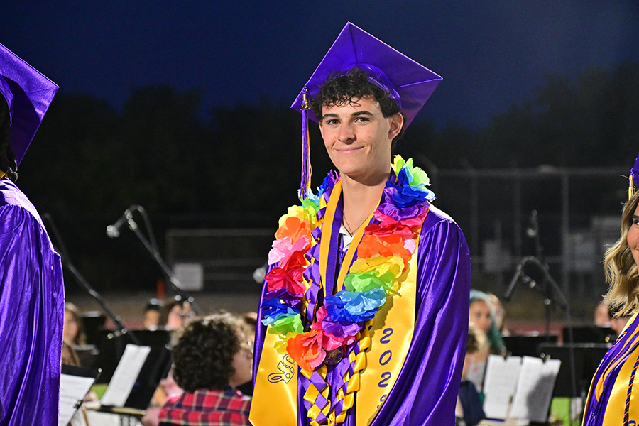 A teen boy in a purple cap and gown and colorful lei smiles on the football field