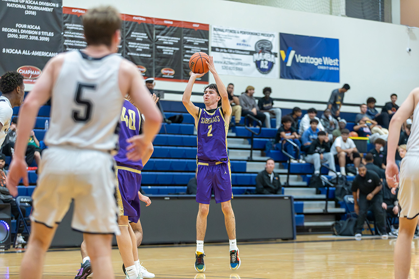 A teen boy in a purple jersey holds the basketball above his head, preparing to shoot a basket