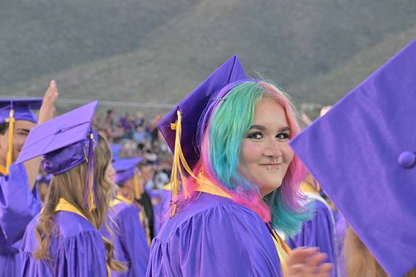 A teen girl with pink and blue hair wearing a purple cap and gown during the graduation ceremony