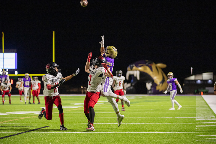 A football player in a purple jersey and gold helmet jumps up to grab the ball as two opposing players run by him