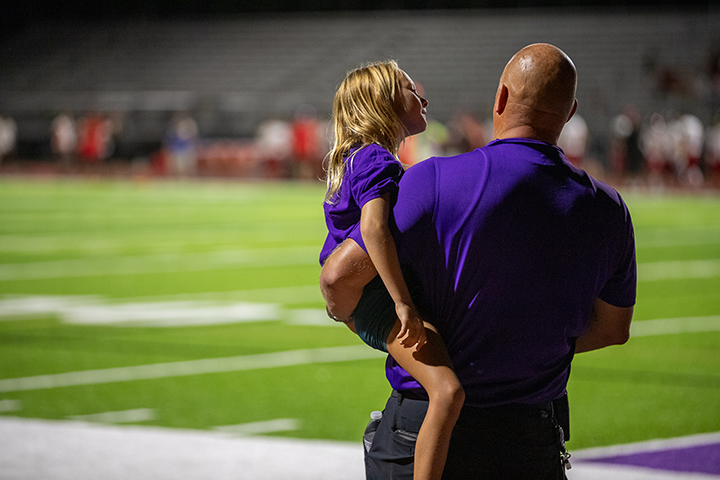 A man in a purple shirt holds a young boy with blonde hair in his arms on a football field