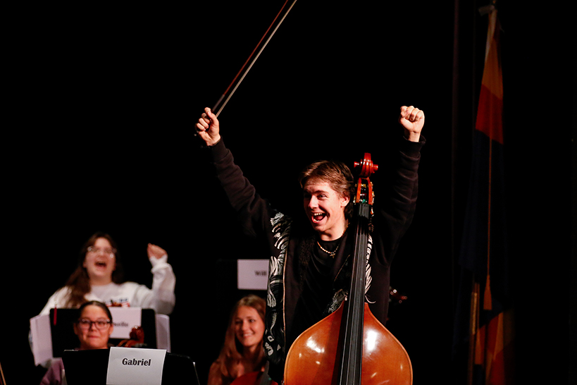 A teen boy playing cello stands up and lifts his bow in excitement