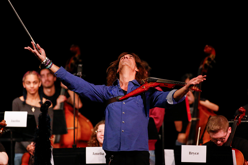 A man lifts his head and arms up triumphantly, while high school musicians play behind him