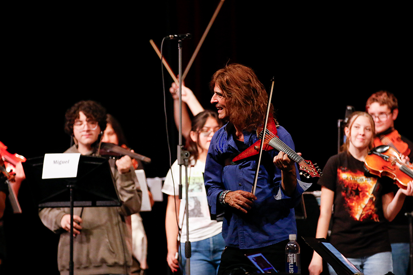 A man playing electric violin smiles at the student musicians around him