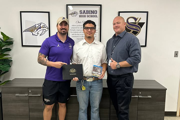 A teen boy in glasses, center, holds up an award with his coach, left, and principal, right