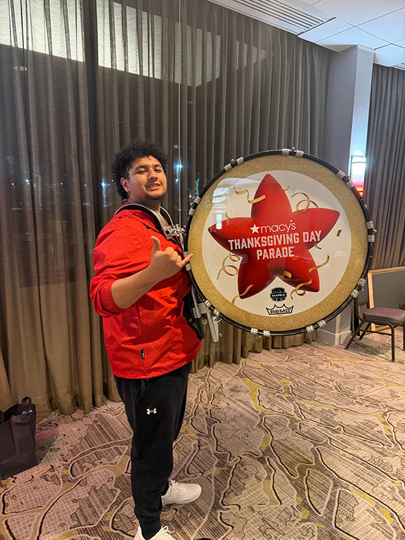 A teen boy in a red jacket gives a thumbs up as he holds his drum