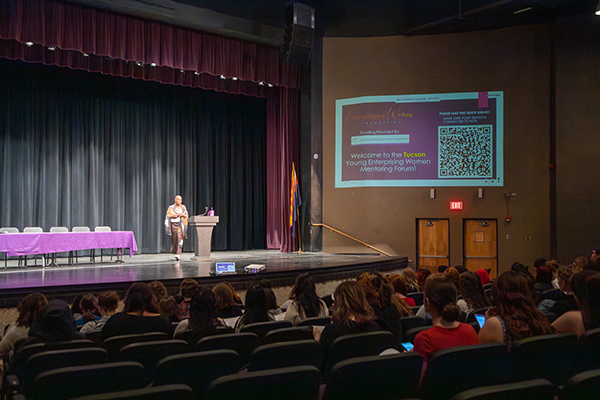 A woman stands at the podium on stage, addressing the group of teen girls