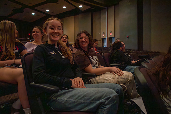 Two teen girls smile in their seats in the auditorium